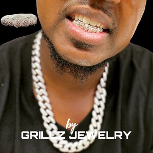 Puede incluir: Primer plano de una persona que lleva una parrilla de oro con detalles de diamantes. La persona tambi&eacute;n lleva un collar grueso de cadena blanca. El texto "by GRILLZ JEWELRY" es visible en la parte inferior de la imagen.
