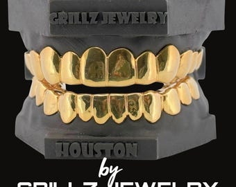 May include: Gold-plated grillz displayed on a black dental mold. The top and bottom grillz are visible, showcasing a full set of teeth. The text "GRILLZ JEWELRY" and "HOUSTON by GRILLZ JEWELRY" are also visible.