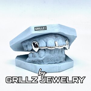 K&ouml;nnte beinhalten: Silberne Grillz f&uuml;r Z&auml;hne, in einer blauen Form dargestellt. Die Grillz sind aus Metall und haben eine gl&auml;nzende Oberfl&auml;che. Der Text "GRILLZ JEWELRY" ist unterhalb der Form sichtbar.