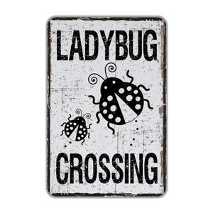 May include: A white metal sign with black text and a black ladybug graphic. The sign reads "Ladybug Crossing".