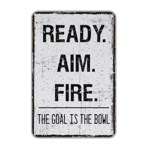 May include: A white metal sign with black text that reads "READY. AIM. FIRE. THE GOAL IS THE BOWL". The sign has a distressed, vintage look.