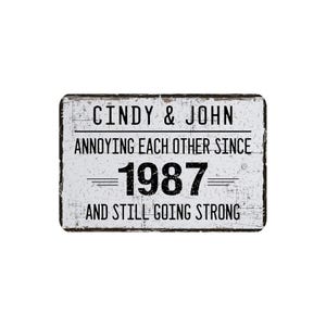 May include: A rectangular, distressed white sign with black text. The sign reads "Cindy & John Annoying Each Other Since 1987 And Still Going Strong." The sign has a black border and a vintage aesthetic.