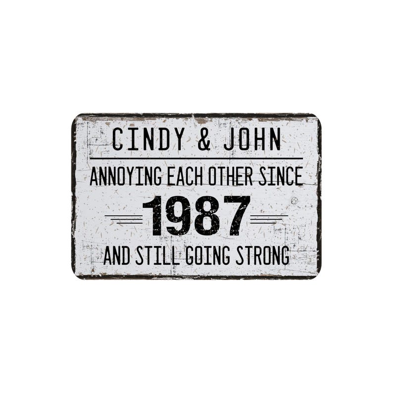 May include: A rectangular, distressed white sign with black text. The sign reads "Cindy & John Annoying Each Other Since 1987 And Still Going Strong." The sign has a black border and a vintage aesthetic.