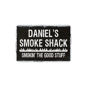 May include: A rectangular black sign with white text that reads "DANIEL'S SMOKE SHACK" above "SMOKIN' THE GOOD STUFF". A white flame design is below the middle text. The sign has a distressed, textured appearance.