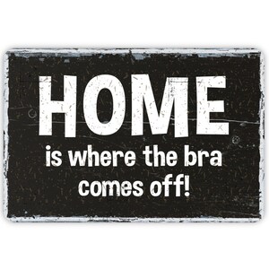 May include: Black and white sign with distressed wood texture. The sign reads "HOME is where the bra comes off!"