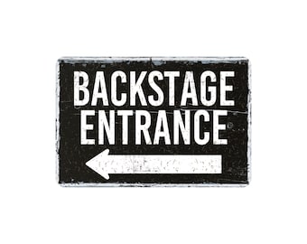 May include: A rectangular black sign with a distressed finish featuring the white text BACKSTAGE ENTRANCE in all capital letters. Below the text is a white horizontal arrow pointing to the left, indicating a direction for authorized personnel.