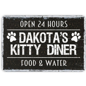 May include: Black and white sign with distressed wood grain texture. The sign reads "Open 24 Hours Dakota's Kitty Diner Food & Water" with paw prints on either side of the name.