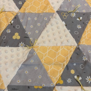 May include: A quilted blanket with a bee and honeycomb theme. The blanket features triangular patches in yellow, gray, and white. Some triangles have bee and honeycomb patterns, while others have daisy patterns. The blanket is tied with yellow yarn.