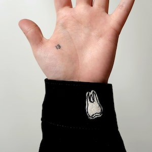 Puede incluir: Puño negro con un diseño de diente bordado en blanco. El número "13" está tatuado en la muñeca. Una mano abierta con los dedos extendidos sobre un fondo blanco.