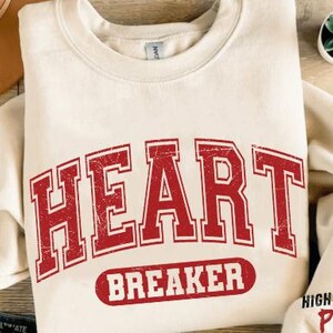 Op de afbeelding: Een crèmekleurig sweatshirt met een rode vintage-stijl grafische afbeelding met de tekst "HEART BREAKER".