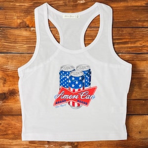 Puede incluir: Camiseta sin mangas blanca con un gráfico de tres latas de cerveza con un diseño de estrellas y rayas azules y blancas. El gráfico incluye el texto "Ameri Can" en rojo y azul.