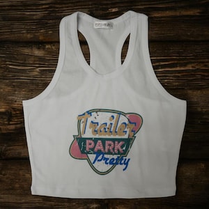 May include: White racerback crop top with the text "Trailer Park Pretty" in a colorful, retro design. The top is made of a soft material and features a scoop neck and a racerback design.