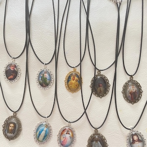Könnte beinhalten: Eine Sammlung religiöser Anhänger-Halsketten. Jede Halskette hat einen ovalen Anhänger mit einem religiösen Bild, der in einem verzierten Metallrahmen gefasst ist. Die Anhänger hängen an schwarzen Kordeln mit silberfarbenen Verschlüssen. Die Bilder zeigen verschiedene religiöse Figuren.