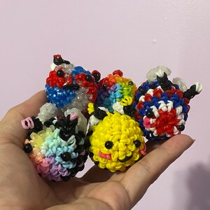 May include: Four colorful rubber band ball toys. The balls are made of rainbow, yellow, red, white, and blue rubber bands. The balls have cute faces with eyes and mouths.