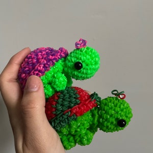 Puede incluir: Dos tortugas de gomas elásticas verdes, una encima de la otra. La tortuga de arriba tiene un caparazón rosa y morado y una cabeza verde. La tortuga de abajo tiene un caparazón rojo y verde y una cabeza verde.