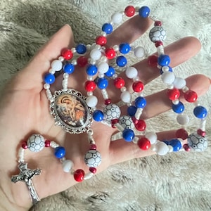 May include: A rosary featuring red, white, and blue beads, including some beads patterned like soccer balls. The rosary includes a silver-toned metal crucifix and an oval centerpiece depicting a religious figure in a decorative silver-toned frame.