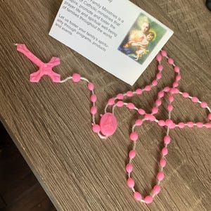 May include: Pink plastic rosary beads with a pink plastic crucifix. The beads are strung on a white string. The crucifix has a white string attached to it. The rosary is on a wooden surface. The image also includes a card with the text "Our Mission Helping Families Pray Holy Cross Family Ministries is a family of Catholic ministries that inspire, promote and foster the prayer life and spiritual well-being of families throughout the world. Let us foster your family's spirituality through programs, products and events."