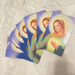 May include: A stack of religious cards featuring a woman with a golden halo and a glowing light emanating from her chest. The woman is wearing a green robe and a yellow head covering. The background is blue with a white and yellow halo.