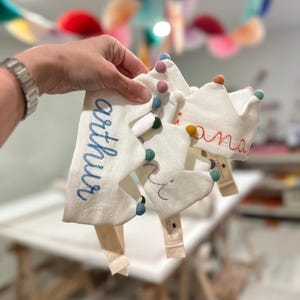 Peut inclure: Trois couronnes en tissu blanc cassé avec des noms brodés dont Arthur, ornées de pompons en feutre colorés sur les pointes. Chaque couronne est dotée d'une sangle élastique beige et d'une petite étiquette en tissu fixée à la base.