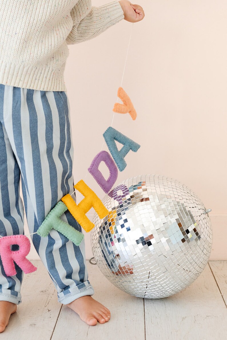 Puede incluir: Un ni&ntilde;o sostiene una pancarta de cumplea&ntilde;os de fieltro con una bola de discoteca en el fondo. La pancarta est&aacute; hecha de letras de fieltro de colores que deletrean "cumplea&ntilde;os".