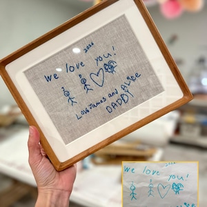 Op de afbeelding: Een rechthoekige houten lijst met een lichtgrijze stoffen inzet met blauwe geborduurde tekst en stokfiguurtekeningen. De tekst luidt: We love you! Love James and Alice DADDY 2026, vergezeld van twee stokfiguren en een hartvormige tekening.