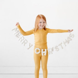 Könnte beinhalten: Eine weiße Filzgirlande mit dem Schriftzug MERRY CHRISTMAS aus einzelnen, ausgestopften Buchstaben, die horizontal gehalten wird. Die Buchstaben sind weich, cremeweiß und durch eine dünne Schnur verbunden, eine festliche Dekoration.