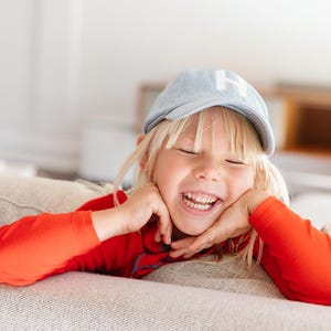May include: A child wearing a pale blue baseball cap with a white "H" and a red long-sleeved top. The child is smiling with eyes closed, resting their chin on their hands.