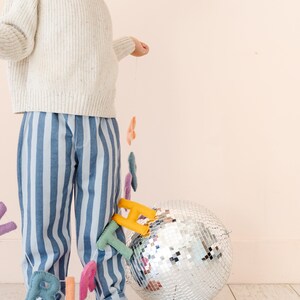 Puede incluir: Un ni&ntilde;o con pantalones a rayas azules y blancas y un su&eacute;ter blanco sostiene una cadena de letras de fieltro que deletrean "cumplea&ntilde;os" en colores arco&iacute;ris. Una bola de discoteca plateada se encuentra en el suelo junto al ni&ntilde;o.