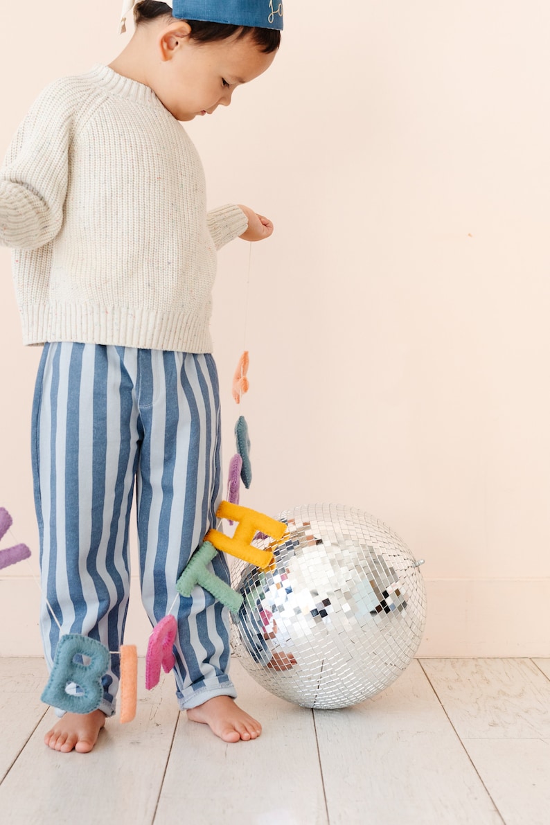 Puede incluir: Un ni&ntilde;o con pantalones a rayas azules y blancas y un su&eacute;ter blanco sostiene una cadena de letras de fieltro que deletrean "cumplea&ntilde;os" en colores arco&iacute;ris. Una bola de discoteca plateada se encuentra en el suelo junto al ni&ntilde;o.