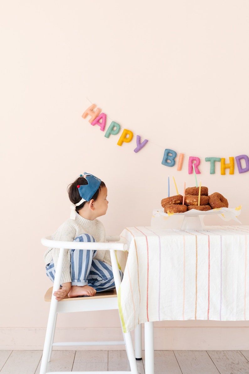 Puede incluir: Un ni&ntilde;o con una corona azul se sienta en una silla blanca y mira una mesa con un plato de donas y velas. La mesa tiene un mantel blanco con rayas de colores. Un cartel con las palabras "Feliz cumplea&ntilde;os" cuelga en la pared detr&aacute;s de la mesa.