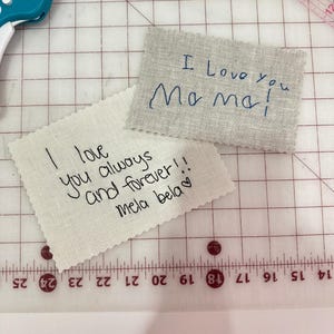 May include: Two rectangular fabric patches with pinked edges featuring embroidered handwriting. One beige patch reads "I love you always and forever!! mela bela" in black thread. The other light gray patch reads "I Love you Mama!" in blue thread.