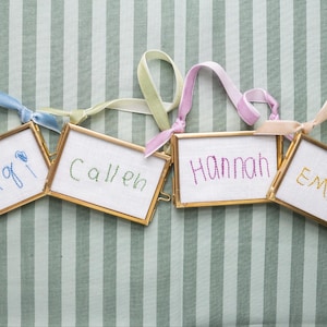 Könnte beinhalten: Vier rechteckige Ornamente mit Messingrahmen und weißen Stoffeinsätzen mit handgestickten Namen. Die Namen Gigi, Callen, Hannah und Emery sind in blauem, grünem, rosa und gelbem Garn gestickt, jeweils mit einem passenden Samtband versehen.