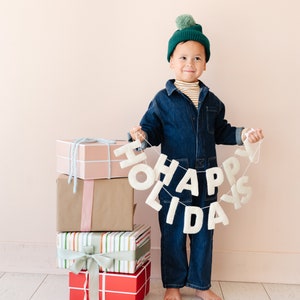 K&ouml;nnte beinhalten: Ein wei&szlig;es Filzbanner mit dem Text HAPPY HOLIDAYS in Gro&szlig;buchstaben, gehalten von einem Kind mit einer dunkelgr&uuml;nen Strickm&uuml;tze mit Bommel, einem gestreiften Oberteil und einem dunkelblauen Jeans-Overall, neben einem Stapel aus vier Geschenkboxen.