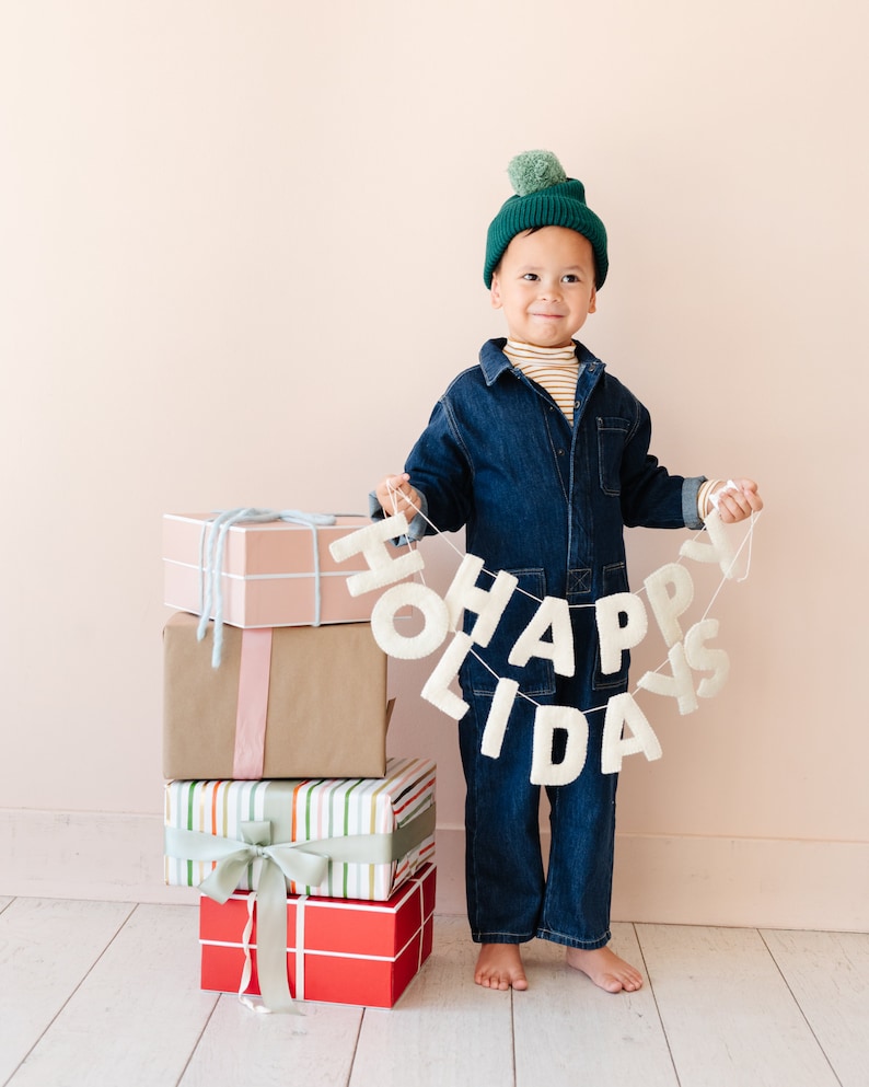 K&ouml;nnte beinhalten: Ein wei&szlig;es Filzbanner mit dem Text HAPPY HOLIDAYS in Gro&szlig;buchstaben, gehalten von einem Kind mit einer dunkelgr&uuml;nen Strickm&uuml;tze mit Bommel, einem gestreiften Oberteil und einem dunkelblauen Jeans-Overall, neben einem Stapel aus vier Geschenkboxen.