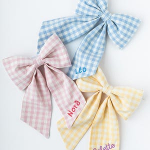 May include: Four large gingham bows in pastel colors: blue, pink, and yellow. Each bow has a long tail and is embroidered with a name: Leo, Nora, and Colette. The bows are arranged on a white background.