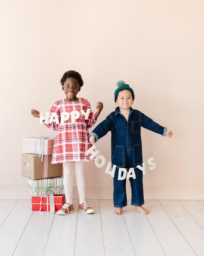 K&ouml;nnte beinhalten: Eine wei&szlig;e Filzgirlande mit dem Text HAPPY HOLIDAYS, gehalten von zwei Kindern. Ein Kind tr&auml;gt ein rot kariertes Kleid mit wei&szlig;er Strumpfhose und Turnschuhen, das andere einen dunkelblauen Jeans-Overall mit einer gr&uuml;nen Bommelm&uuml;tze.