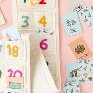 Könnte beinhalten: Cremefarbener Adventskalender aus Filz mit bunten, gestickten Zahlen und kleinen Taschen. Enthält Karten mit Illustrationen und Text wie "Cut Out Paper Snowflakes" und "Christmas Bucket List". Ein Lutscher befindet sich in einer Tasche.