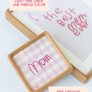 May include: A framed embroidery with the text "the best mom ever" in red thread. A square coaster with "Mom" embroidered in pink thread on pink gingham fabric sits below. Both items have wooden frames.