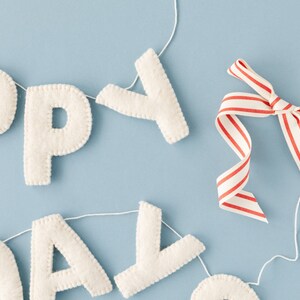 K&ouml;nnte beinhalten: Eine Filzbuchstaben-Girlande, die HAPPY BIRTHDAY buchstabiert, bestehend aus cremewei&szlig;en, gen&auml;hten Stoffbuchstaben an einer d&uuml;nnen wei&szlig;en Schnur. Ein Zierband mit d&uuml;nnen roten und wei&szlig;en vertikalen Streifen ist nahe dem Y an der Schnur befestigt.