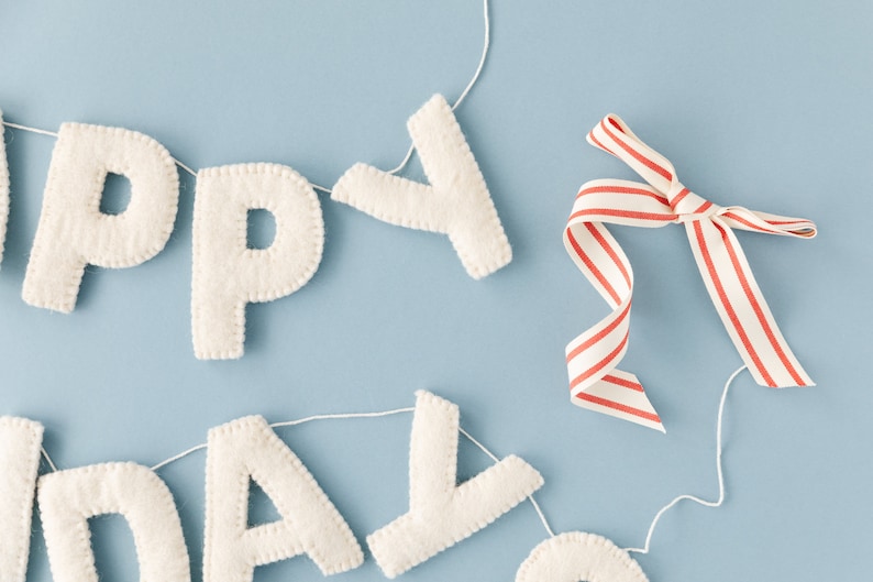 K&ouml;nnte beinhalten: Eine Filzbuchstaben-Girlande, die HAPPY BIRTHDAY buchstabiert, bestehend aus cremewei&szlig;en, gen&auml;hten Stoffbuchstaben an einer d&uuml;nnen wei&szlig;en Schnur. Ein Zierband mit d&uuml;nnen roten und wei&szlig;en vertikalen Streifen ist nahe dem Y an der Schnur befestigt.