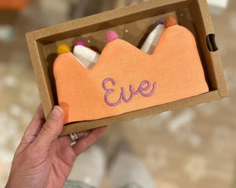 Op de afbeelding: Een perzikkleurige stoffen kroon met de naam Eve geborduurd in paarse draad. De kroon heeft een geschulpte bovenrand versierd met kleine, veelkleurige vilten pompons in geel, roze en bruin, gepresenteerd in een rechthoekige kartonnen doos.