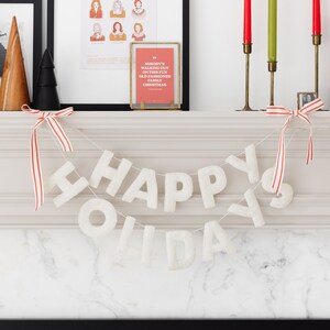 K&ouml;nnte beinhalten: Eine wei&szlig;e Filzbuchstaben-Girlande, die in zwei Reihen HAPPY HOLIDAYS buchstabiert, aufgeh&auml;ngt an einer d&uuml;nnen Schnur mit rot-wei&szlig; gestreiften Schleifen an beiden Enden. Die Buchstaben haben eine weiche, strukturierte Optik vor dem Kaminsims.