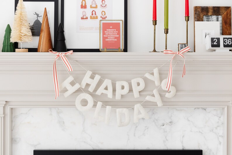 K&ouml;nnte beinhalten: Eine wei&szlig;e Filzbuchstaben-Girlande, die in zwei Reihen HAPPY HOLIDAYS buchstabiert, aufgeh&auml;ngt an einer d&uuml;nnen Schnur mit rot-wei&szlig; gestreiften Schleifen an beiden Enden. Die Buchstaben haben eine weiche, strukturierte Optik vor dem Kaminsims.