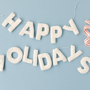 K&ouml;nnte beinhalten: Ein dekoratives Banner mit cremewei&szlig;en, filzartigen Stoffbuchstaben, die den Schriftzug HAPPY HOLIDAYS bilden. Die Buchstaben sind auf einer d&uuml;nnen wei&szlig;en Schnur aufgereiht, und an der rechten Seite ist ein rot-wei&szlig; gestreiftes Band befestigt.