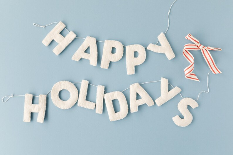 K&ouml;nnte beinhalten: Ein dekoratives Banner mit cremewei&szlig;en, filzartigen Stoffbuchstaben, die den Schriftzug HAPPY HOLIDAYS bilden. Die Buchstaben sind auf einer d&uuml;nnen wei&szlig;en Schnur aufgereiht, und an der rechten Seite ist ein rot-wei&szlig; gestreiftes Band befestigt.