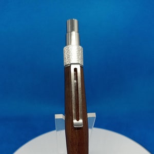 May include: A mechanical pencil with a dark brown wooden barrel and silver metal accents. The pencil features a pocket clip and textured grip sections. The pencil is displayed on a clear acrylic stand against a blue background.
