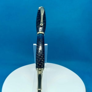 Puede incluir: Un bolígrafo artesanal con un cuerpo estampado en azul oscuro y negro y detalles dorados. El bolígrafo presenta un clip plateado y un diseño elegante y moderno. El bolígrafo se exhibe sobre un soporte blanco sobre un fondo azul.