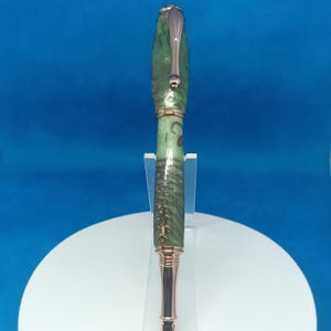 Puede incluir: Un bolígrafo artesanal con un cuerpo veteado verde y marrón y detalles color cobre. El bolígrafo presenta un diseño elegante con un clip y una punta detallada. El bolígrafo se exhibe sobre un soporte blanco sobre un fondo azul.