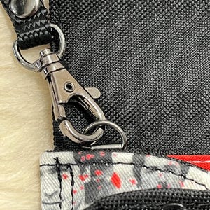 May include: A black fabric bag featuring a white and gray pattern with red paint splatter accents. The bag includes a black nylon strap attached with a silver-toned metal lobster clasp and a black zipper closure on the front pocket section.
