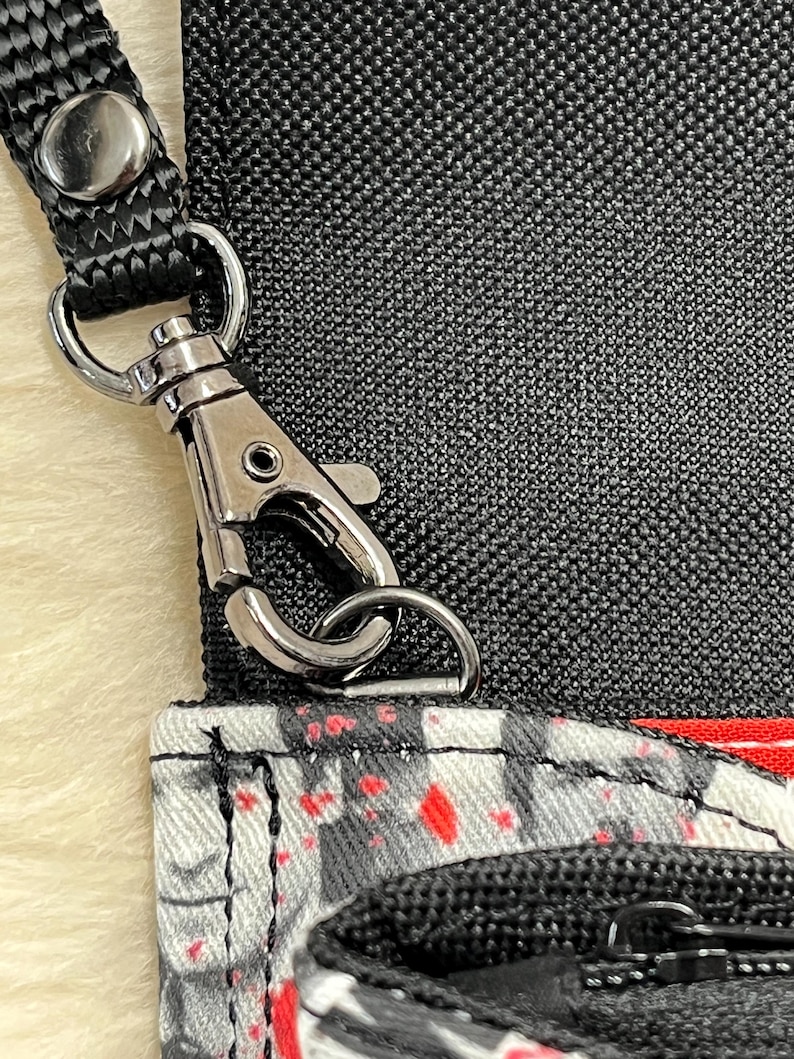May include: A black fabric bag featuring a white and gray pattern with red paint splatter accents. The bag includes a black nylon strap attached with a silver-toned metal lobster clasp and a black zipper closure on the front pocket section.